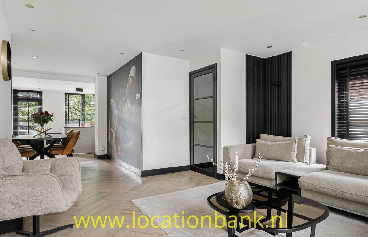 Modernes Wohnzimmer mit beigefarbenen Sofas, rundem Couchtisch, Holzboden und großen Fenstern mit Jalousien.