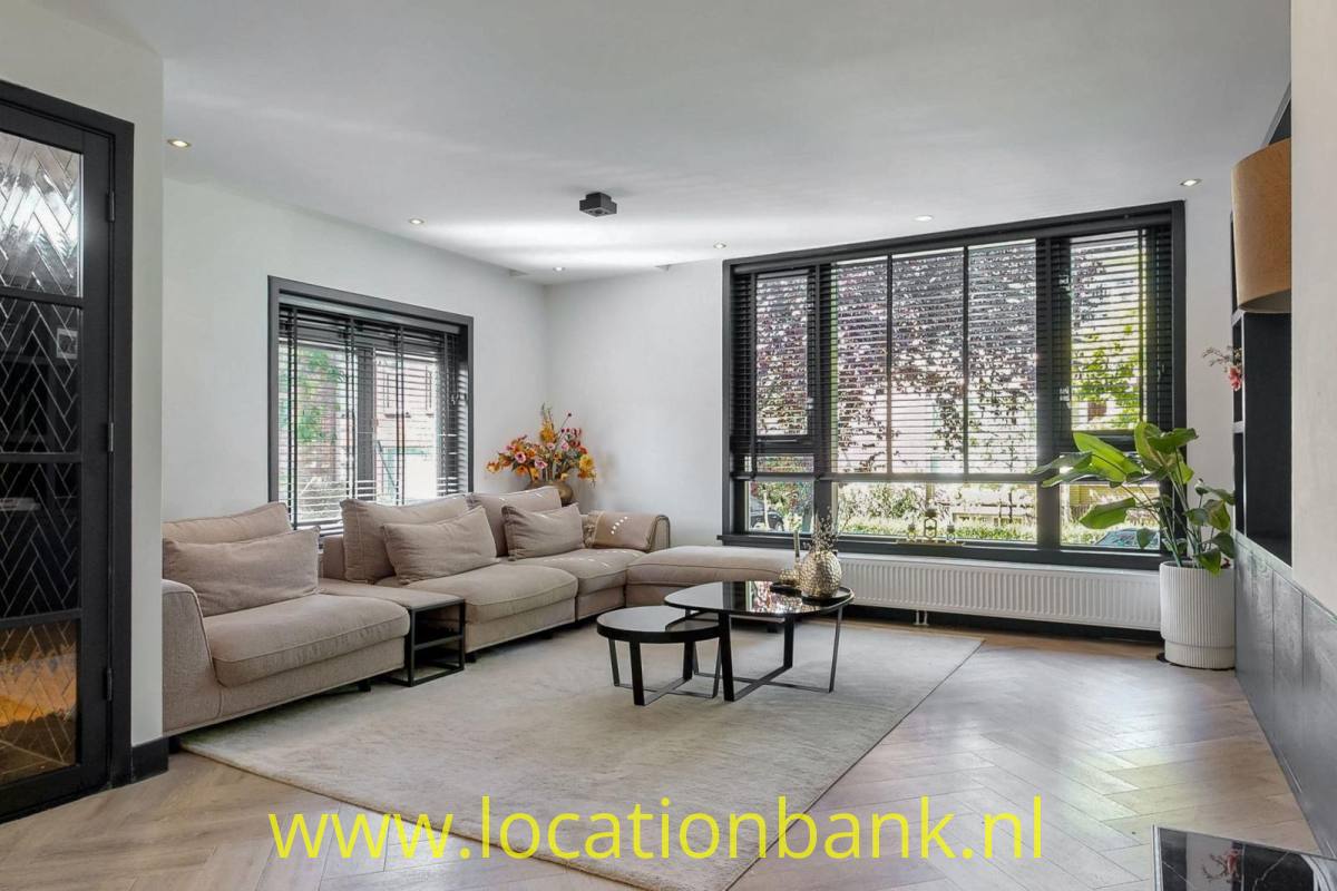 Wohnzimmer mit einem beigefarbenen Ecksofa, runden Couchtischen und großen schwarzen Fenstern mit Blick ins Grüne.