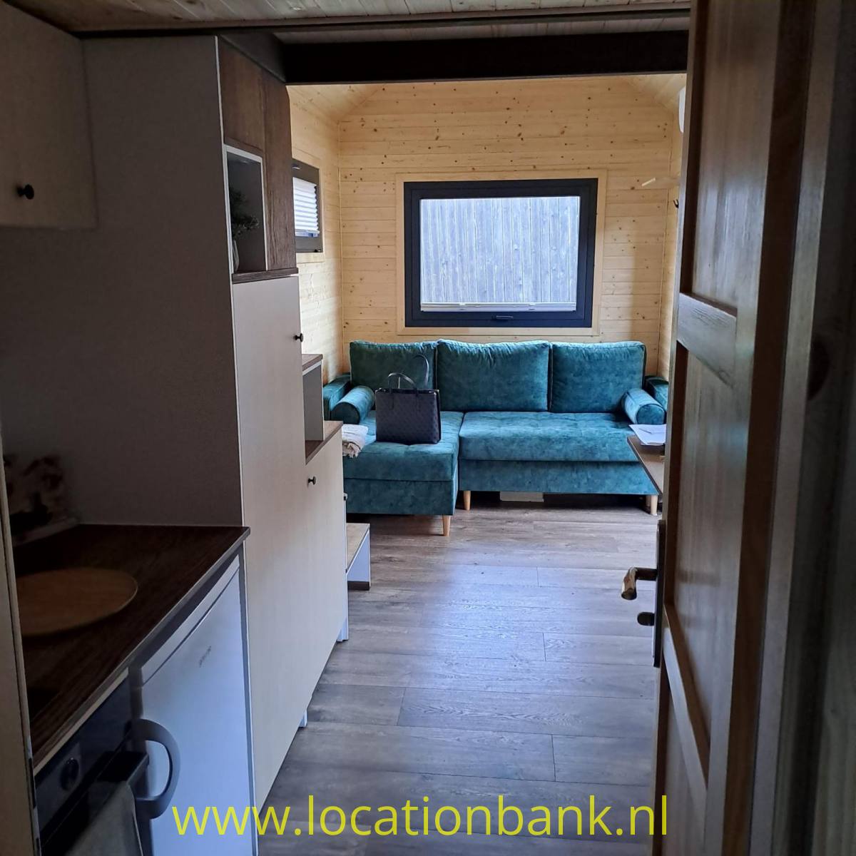 Ein blaugrünes Sofa steht an einer Holzwand unter einem Fenster, sichtbar durch eine offene Tür.