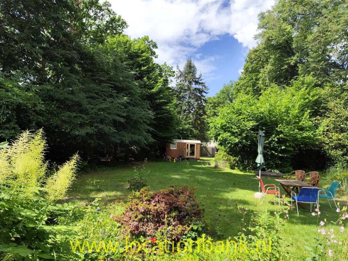 Ein grüner Garten mit hohen Bäumen, blühenden Pflanzen, einem hölzernen Gartenhaus und einer Sitzecke mit Sonnenschirm.