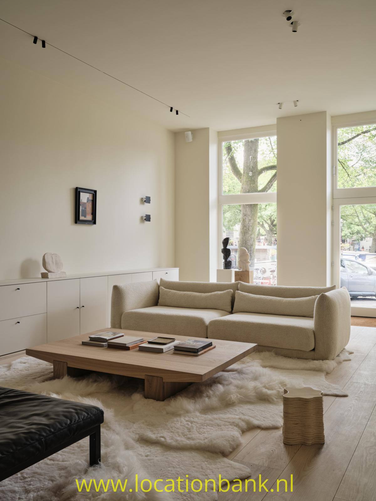 Stimmungsvolles, geräumiges Wohnzimmer mit einer sanften, natürlichen Ausstrahlung. Ein großzügiges helles Sofa, ein niedriger Couchtisch aus Holz, Wollteppiche und dezente Kunstobjekte verleihen dem Raum eine zeitgemäße, ruhige Atmosphäre. Große Fenster sorgen für viel Tageslicht und bieten einen Blick auf die Straße und das Grün im Außenbereich.