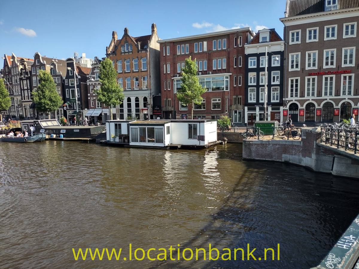 Ein Hausboot liegt in einem Kanal, dahinter stehen schmale hohe amsterdam Häuser und eine steinerne Kaimauer.