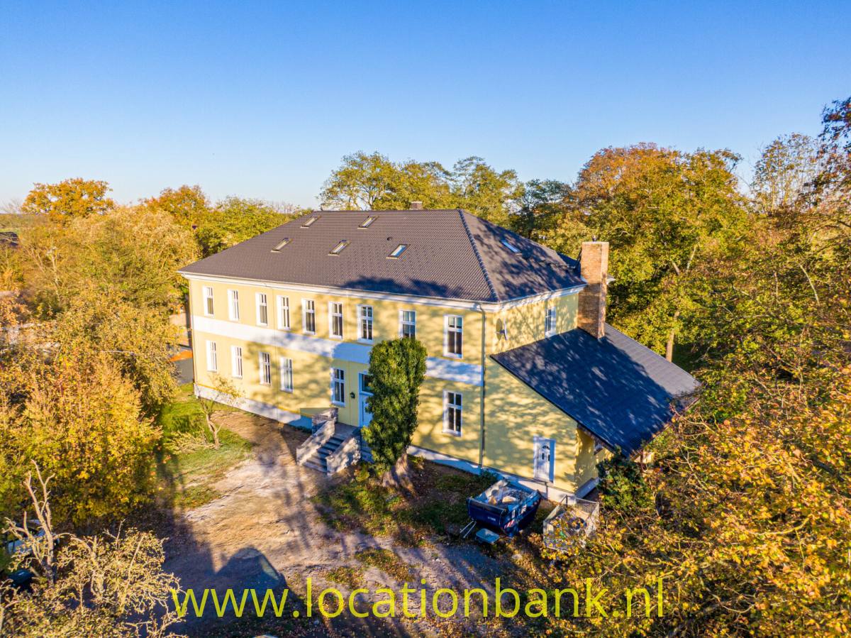 Großes gelbes zweigeschossiges Haus mit dunklem Dach und weißen Fensterrahmen, umgeben von herbstlich gefärbten Bäumen.
