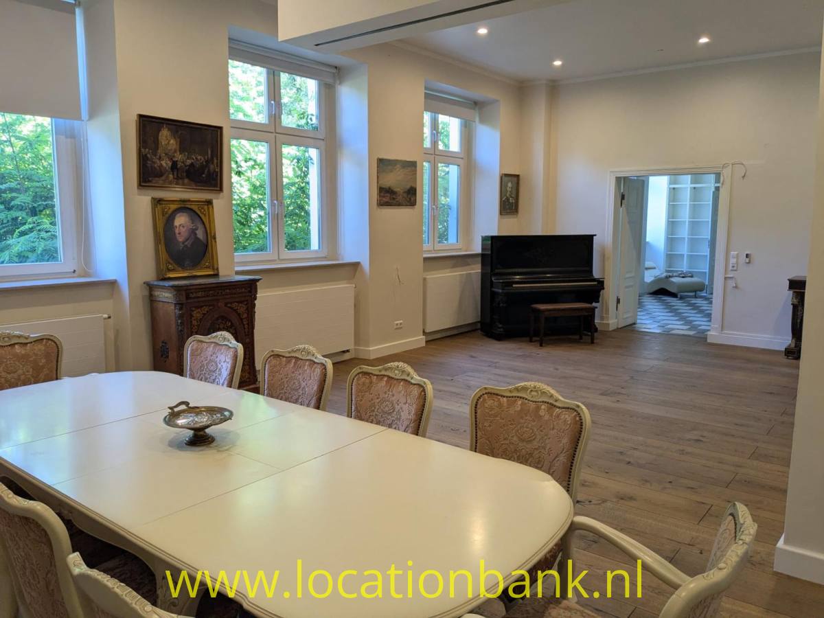 Geräumiges Esszimmer mit einem großen weißen Tisch, mehreren Stühlen, einem schwarzen Klavier und gerahmten Porträts.