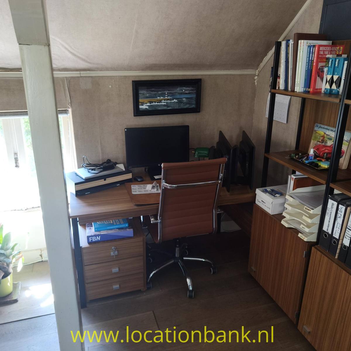 Ein Arbeitsplatz mit Schreibtisch, Computermonitor, Tastatur, Bürostuhl und einem mit Büchern gefüllten Regal.