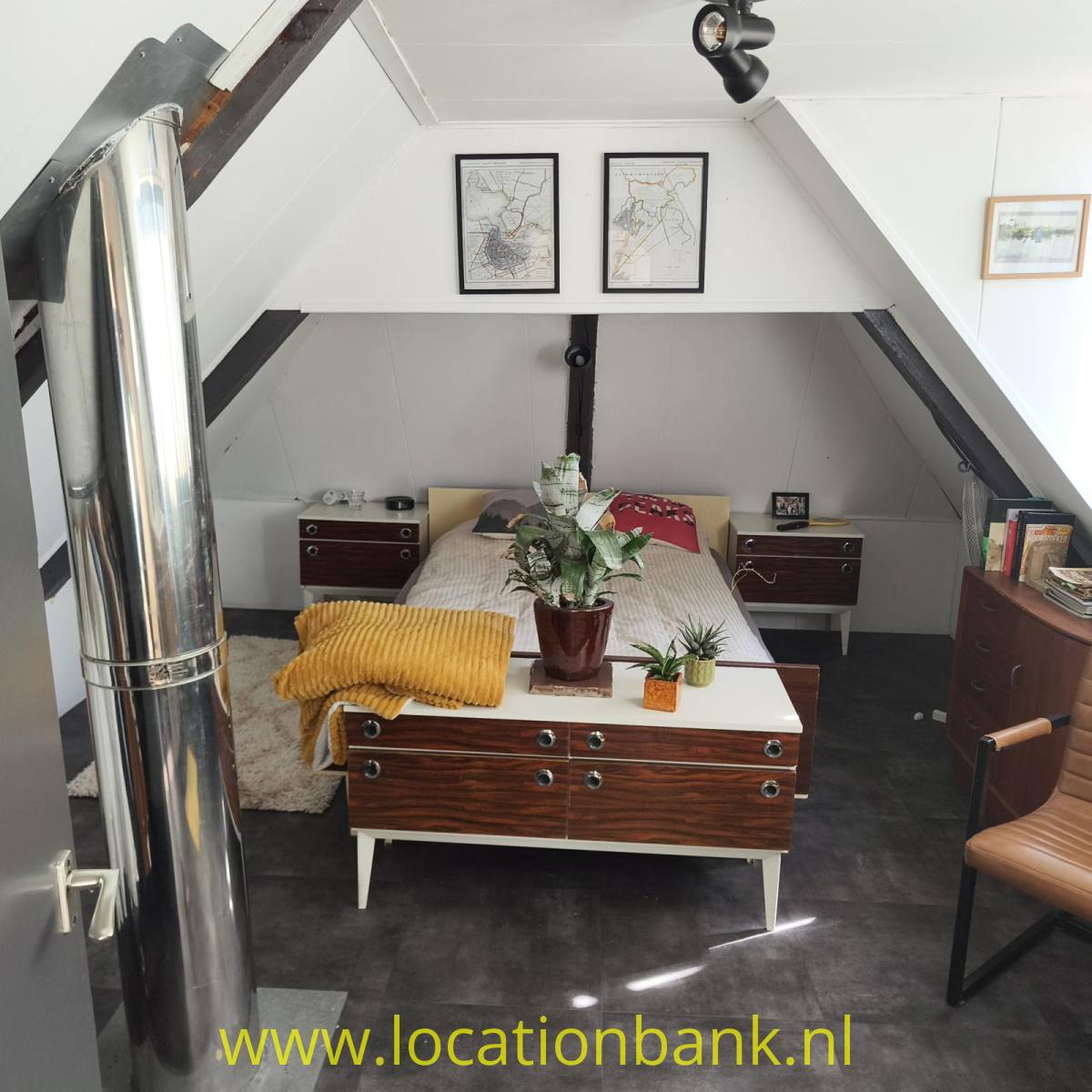 Dachboden Schlafzimmer mit schräger Decke, hölzerner Kommode, Bett, Pflanzen und einem auffälligen Metallofenrohr.