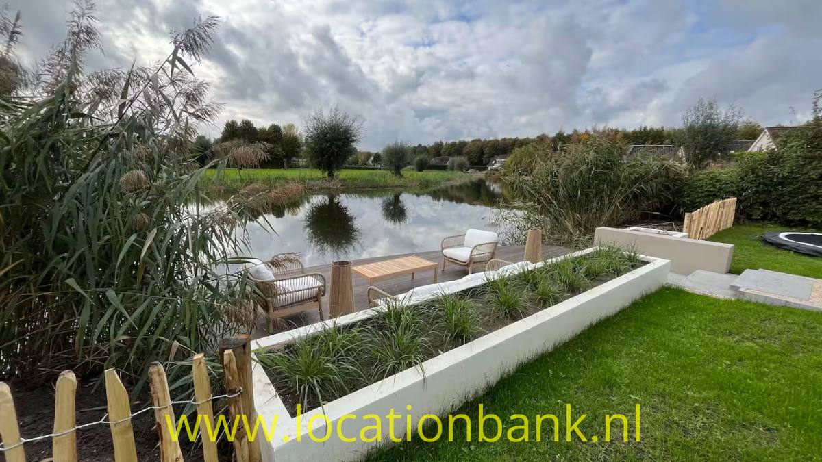 Rasen und erhöhte, weiß eingefasste Pflanzbeete an einem Teich sowie eine Holzterrasse mit Stühlen und hohem Schilf.