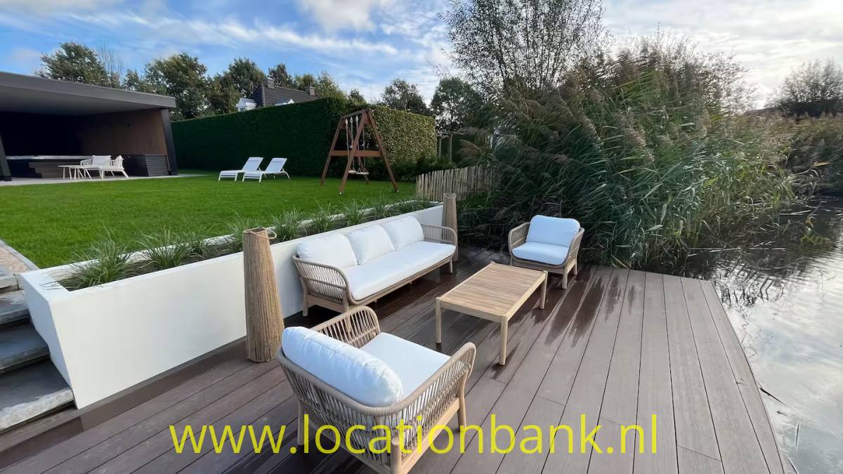 Eine Holzterrasse mit einem geflochtenen Sofa, zwei Sesseln und einem niedrigen Tisch neben einem Teich.