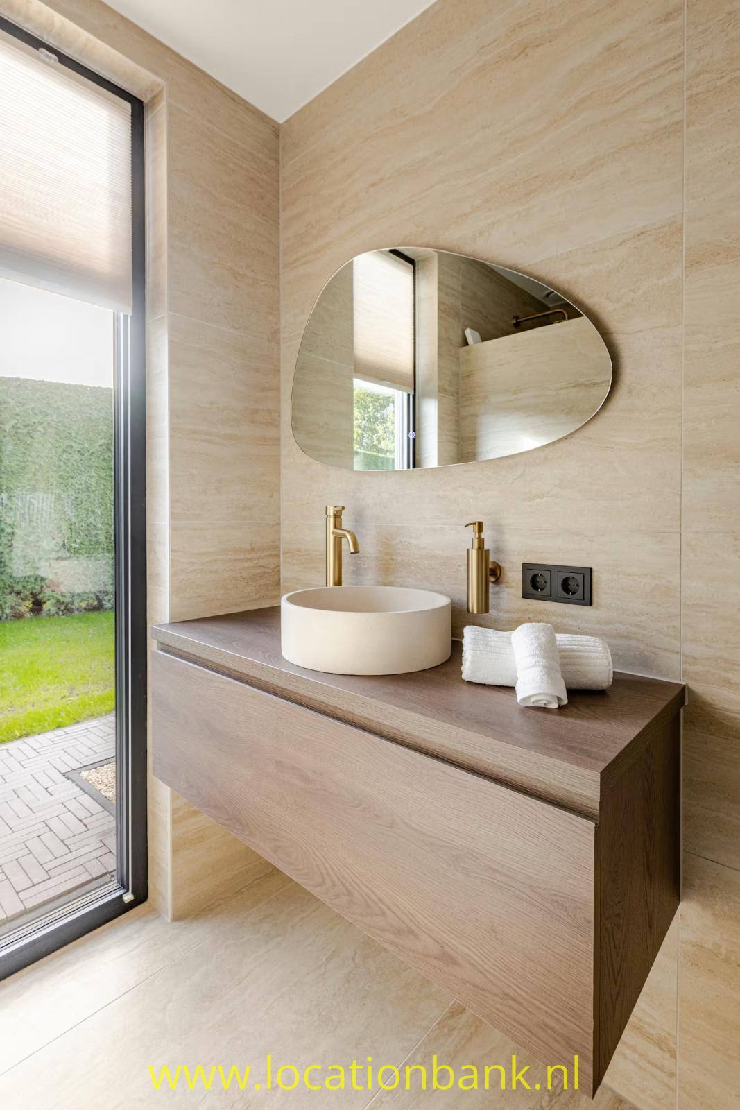 Waschtisch aus Holz mit rundem Waschbecken, goldfarbenen Armaturen, Handtüchern und einem organisch geformten Spiegel neben einem Fenster.