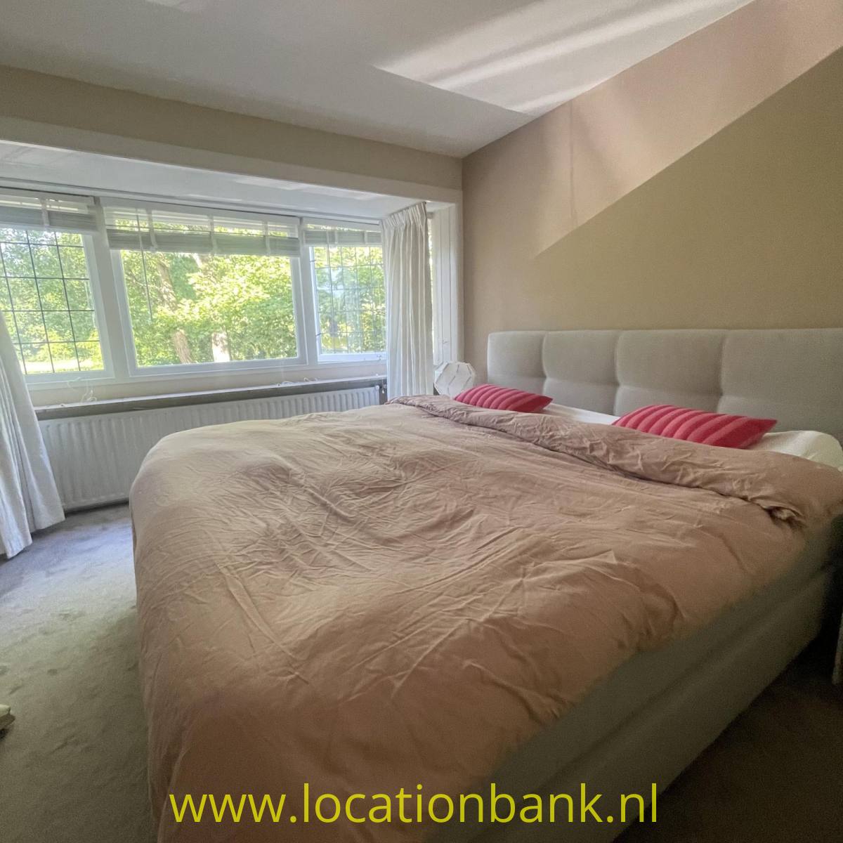 Ein großes Bett mit einer beigen Bettdecke, rosafarbenen Kissen und einem gepolsterten Kopfteil steht vor einem großen Fenster.