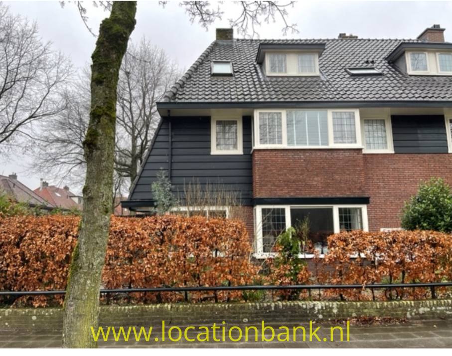Haus mit dunkler Fassadenverkleidung, einer Backsteinfassade, Gauben, Dachfenstern, einer Hecke und einem Baum.