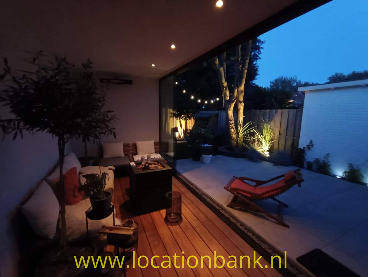Überdachte Terrasse mit Sitzbereich, Holzdeck, Pflanzen und roten Liegestühlen neben einem gepflasterten Innenhof.
