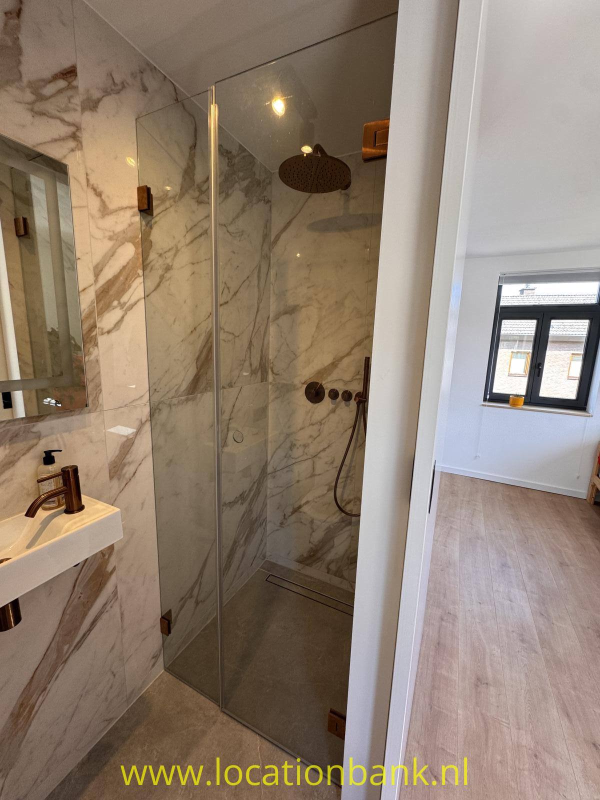 Badezimmer mit einer gläsernen Duschkabine, Wandfliesen in Marmoroptik und einem Waschbecken neben einem angrenzenden Raum.