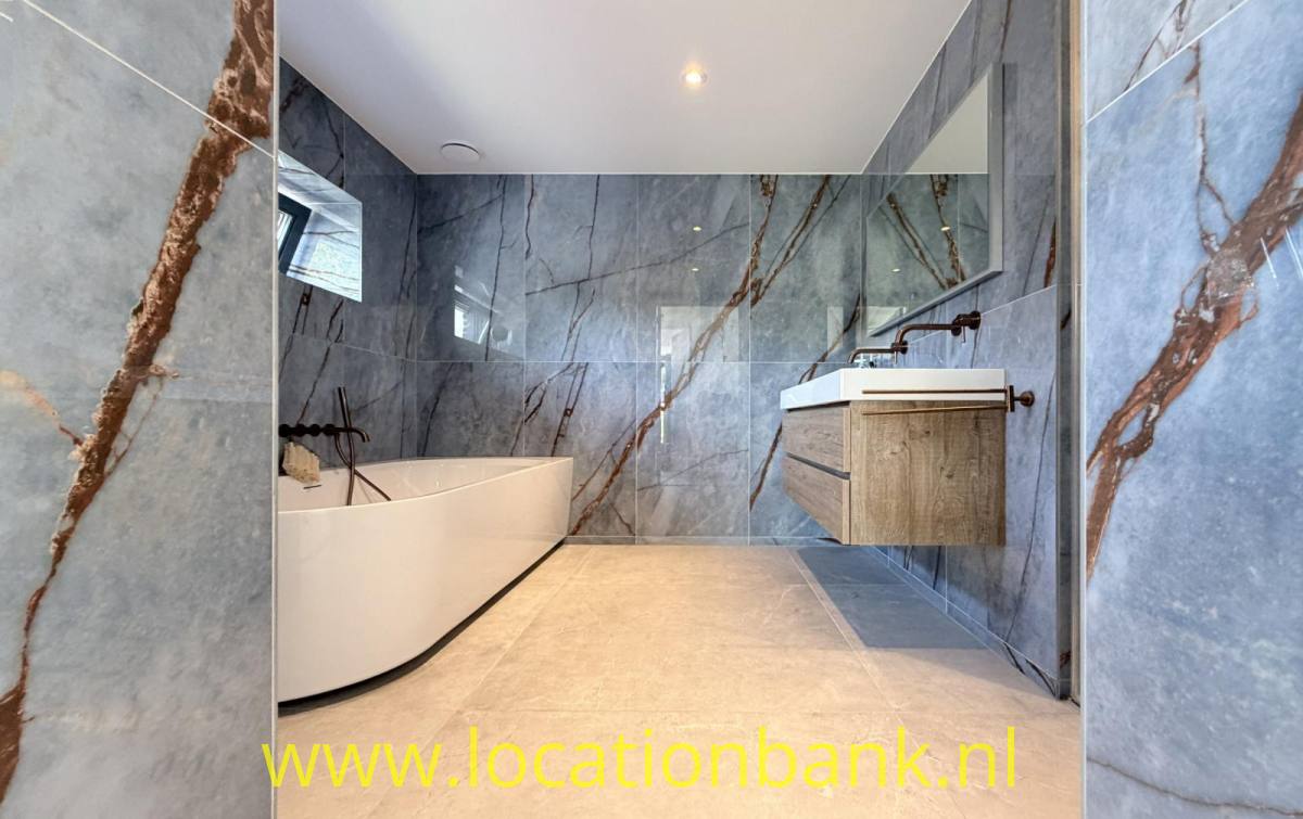 Badezimmer mit einer freistehenden weißen Badewanne, einem hölzernen Waschtischunterschrank und grauen Wandfliesen mit braunen Adern.