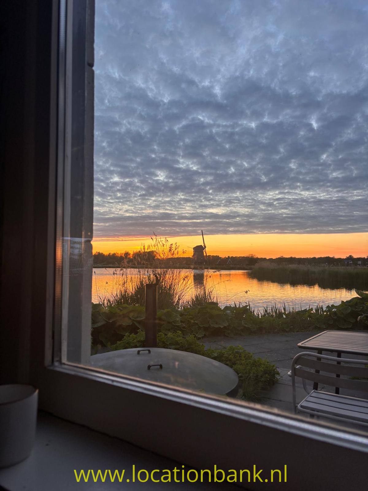 Ein von Schilf gesäumter See spiegelt den orangefarbenen Sonnenuntergang, gesehen durch ein Fenster.
