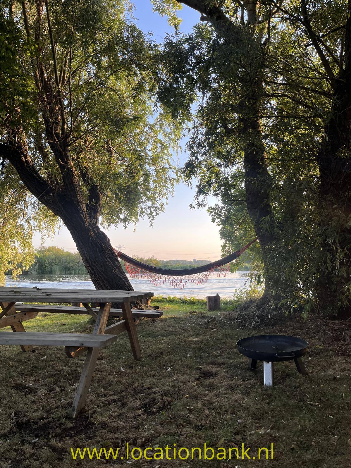 Ein hölzerner Picknicktisch steht unter Bäumen im Garten neben einer Hängematte am Wasser.