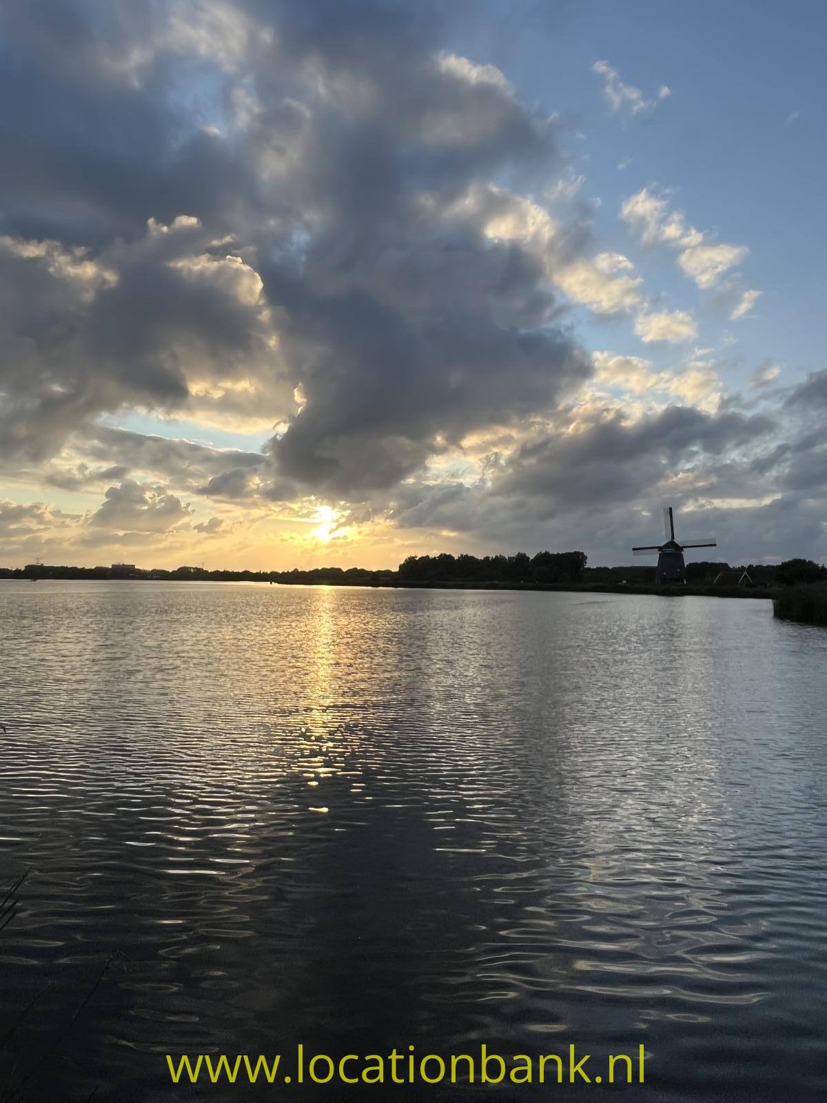 Eine Windmühle steht am Wasser, während die tief stehende Sonne durch dunkle Wolken scheint.