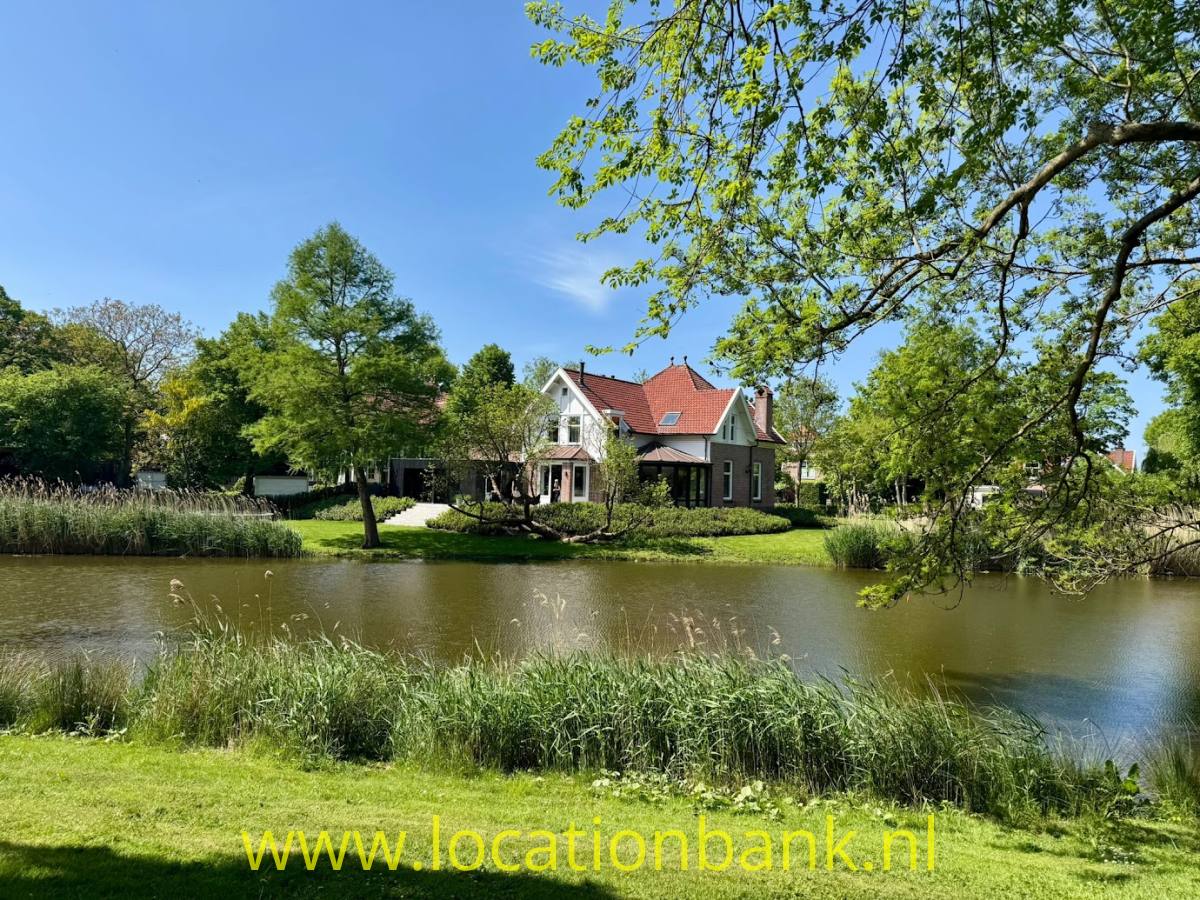 Ein Haus mit roten Dächern liegt hinter einem Teich, umgeben von Bäumen und Schilf.