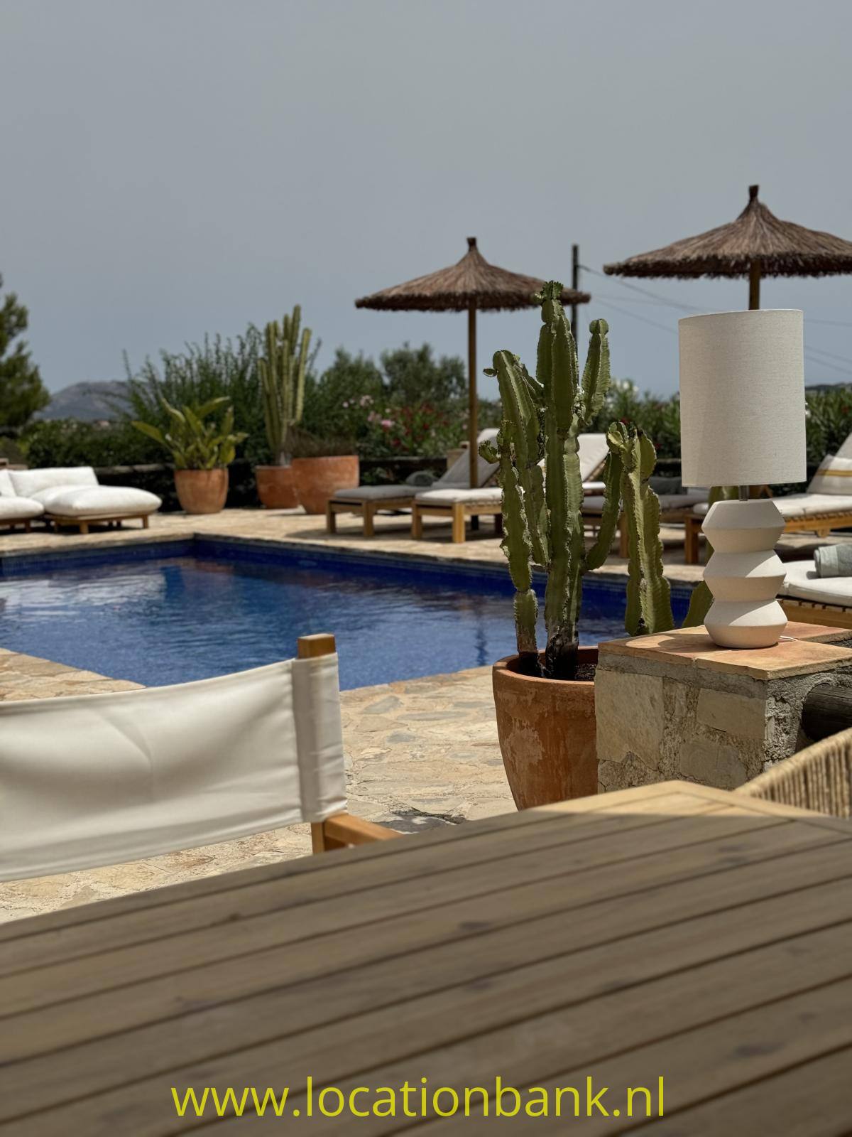 Swimmingpool mit Liegen, Sonnenschirmen, Kakteen, einer Außenleuchte und einer Holzterrasse im Vordergrund.