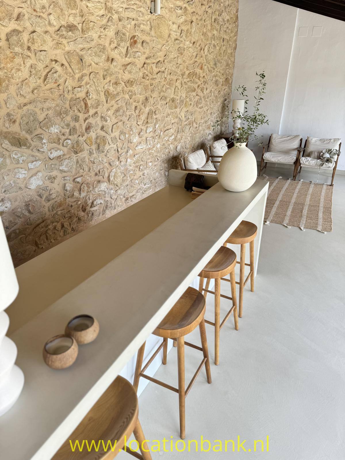 Lange beigefarbene Theke mit Holzstühlen, einer großen Vase und einer Natursteinwand.