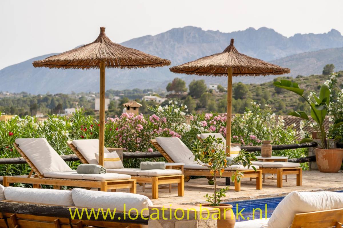Sonnenliegen und Strohs­­chirme stehen zwischen blühenden Pflanzen neben einem kleinen Pool, im Hintergrund sind Berge zu sehen.