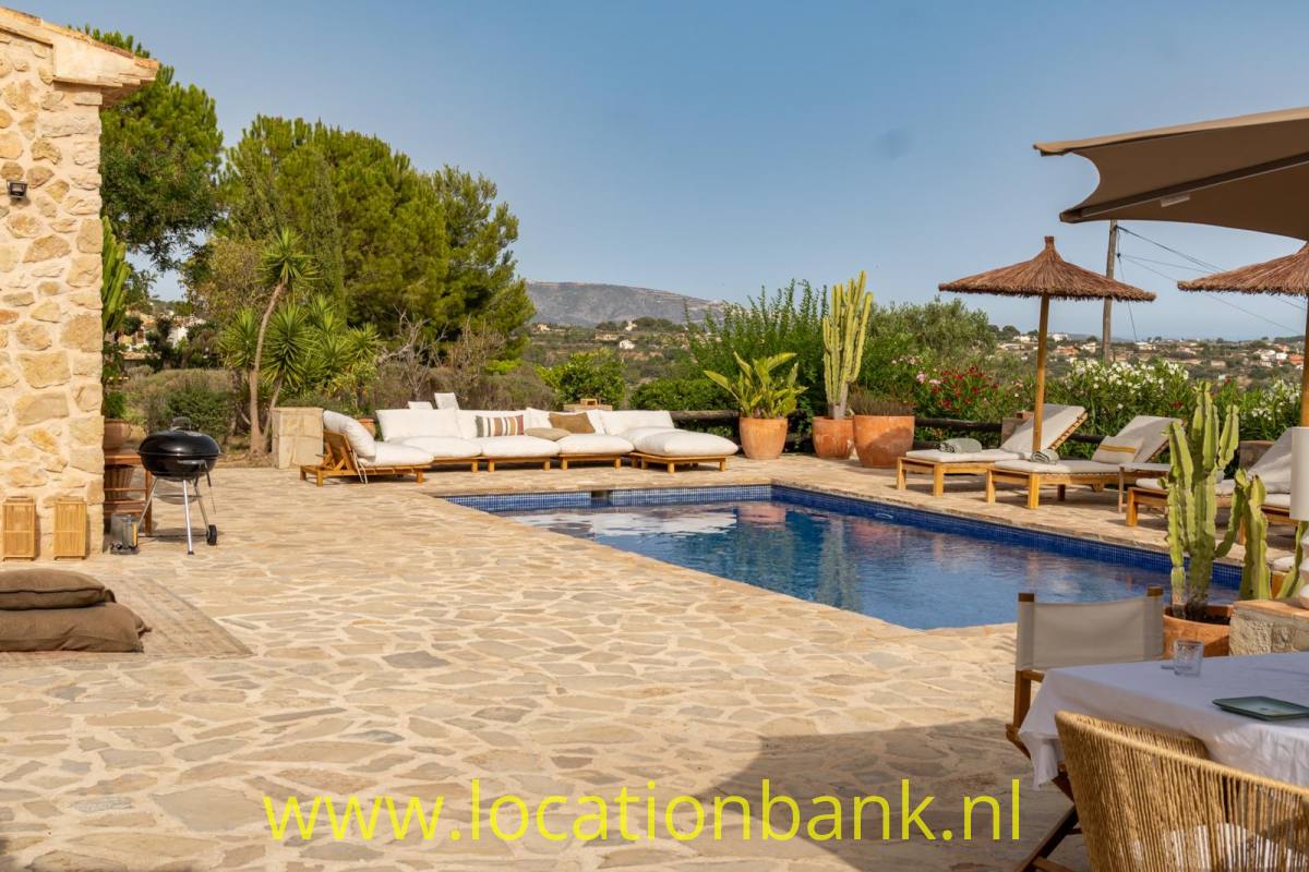 Swimmingpool auf einer Steinterrasse mit Liegestühlen, Sonnenschirmen und Blick auf bewaldete Hügel.