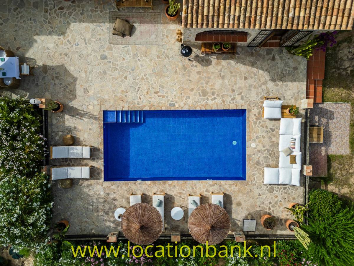 Rechteckiger blauer Swimmingpool in einem mit Naturstein gepflasterten Innenhof, umgeben von Liegestühlen, Sonnenschirmen und weißen Sitzmöbeln.