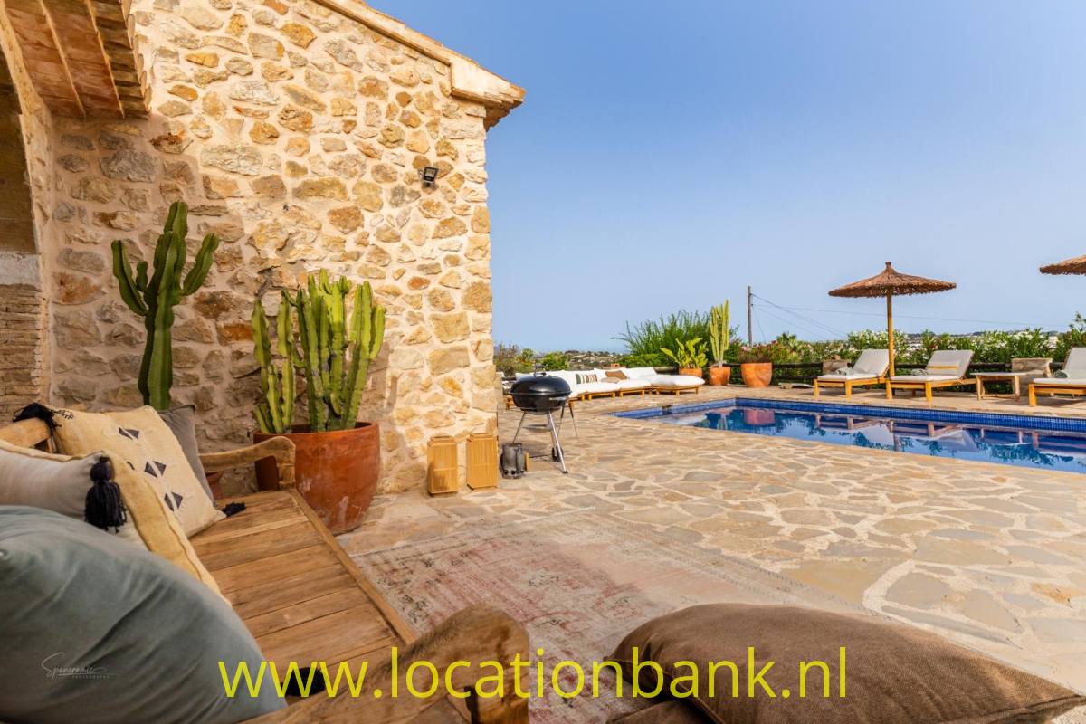 Steinterrasse neben einem Steingebäude mit Swimmingpool, Liegestühlen, Sonnenschirmen und Topfpflanzen.