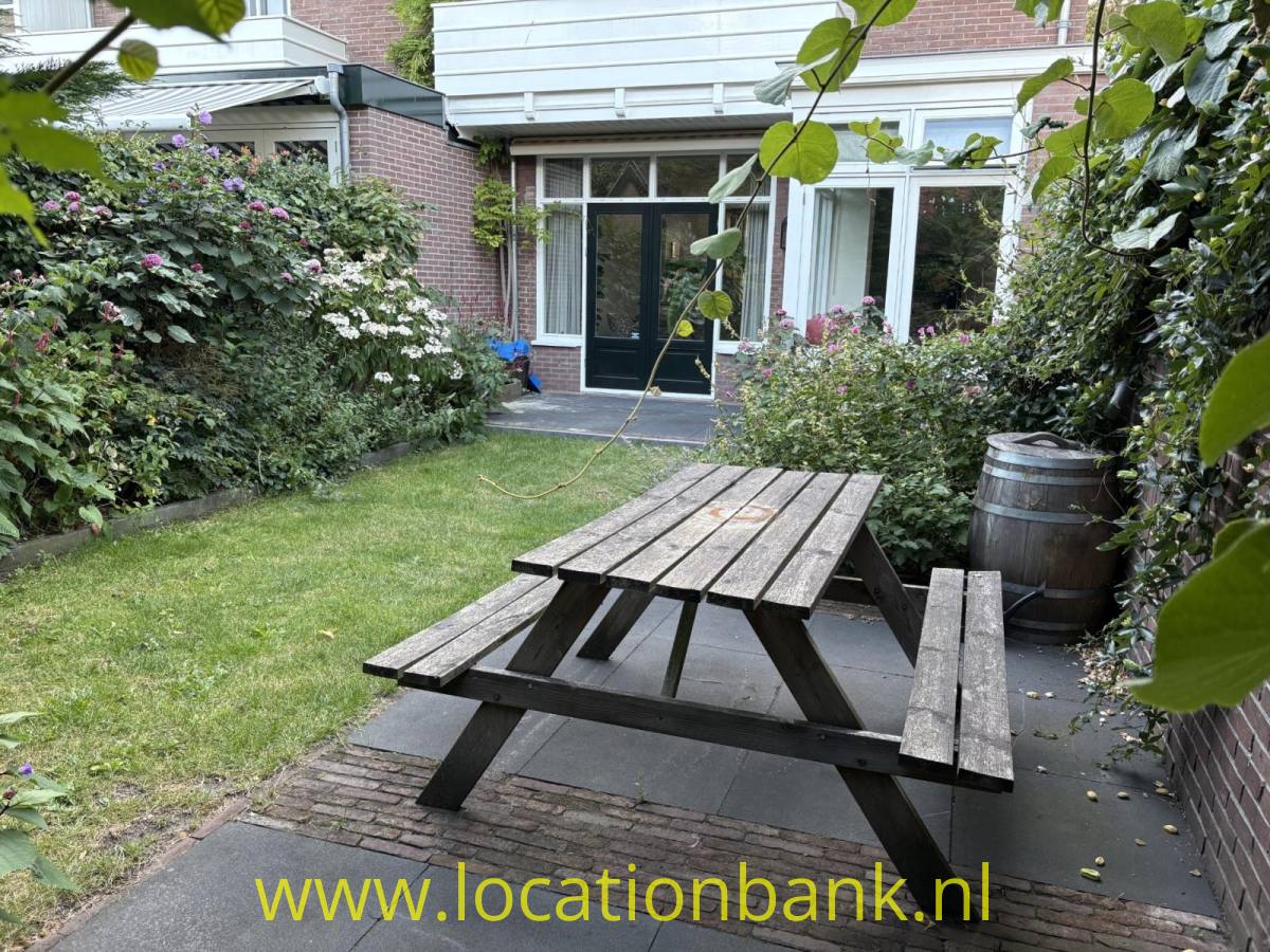 Ein hölzerner Picknicktisch steht auf einer gepflasterten Terrasse in einem grünen Innenhof neben einem Ziegelgebäude.