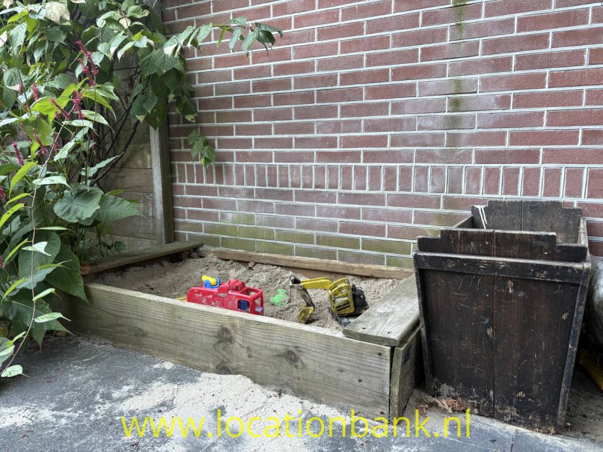Sandbak buntem Spielzeug gefüllte Holzkiste steht vor einer Backsteinmauer neben einem dunklen Holzbehälter.