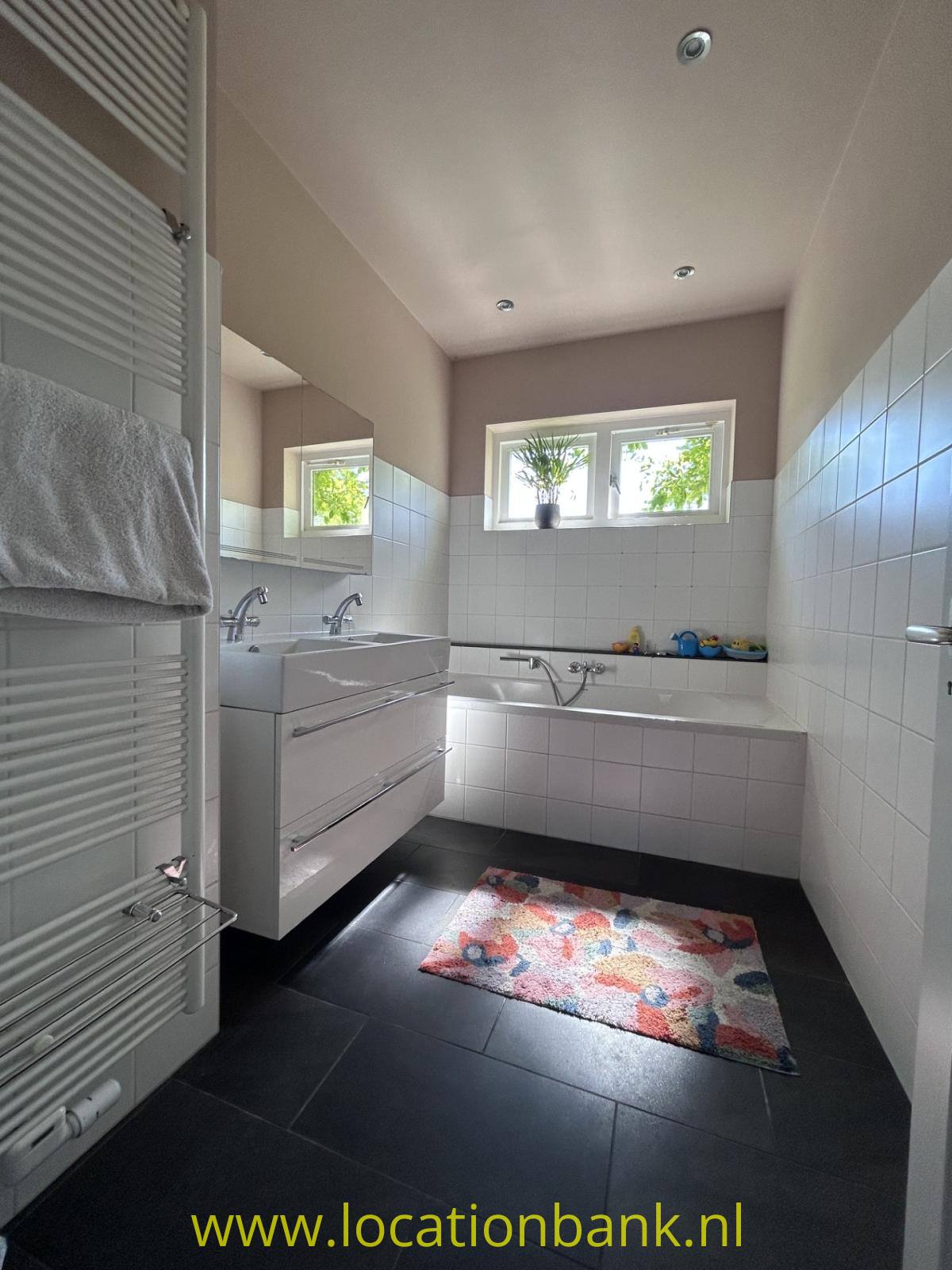 Badezimmer mit weiße Wandfliesen, eine Badewanne, einen Waschtischunterschrank, ein Fenster und einen bunten Teppich.