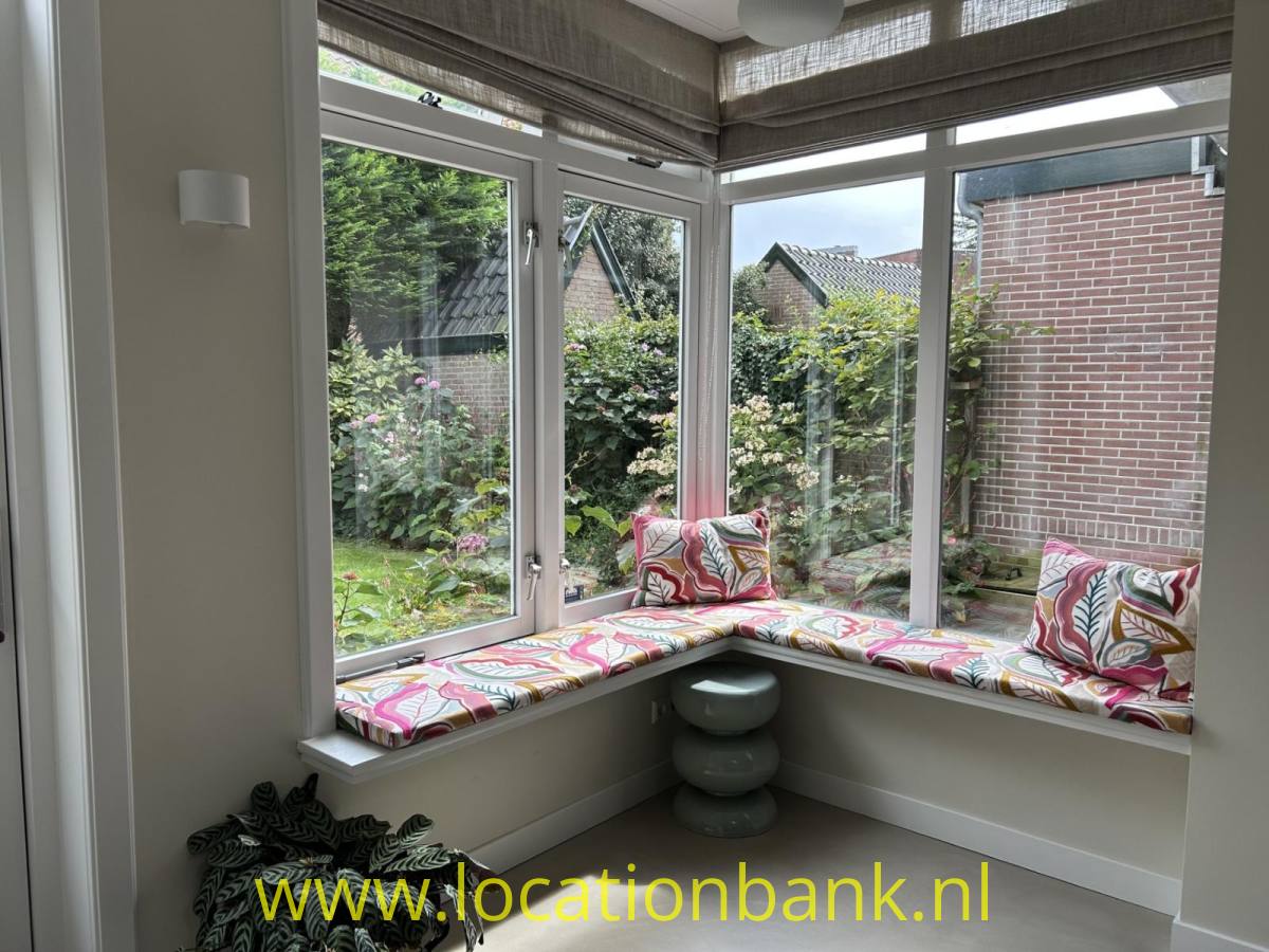 Ein Eckfenster mit gepolsterter Fensterbank und gemusterten Kissen bietet einen Blick auf einen grünen Garten.