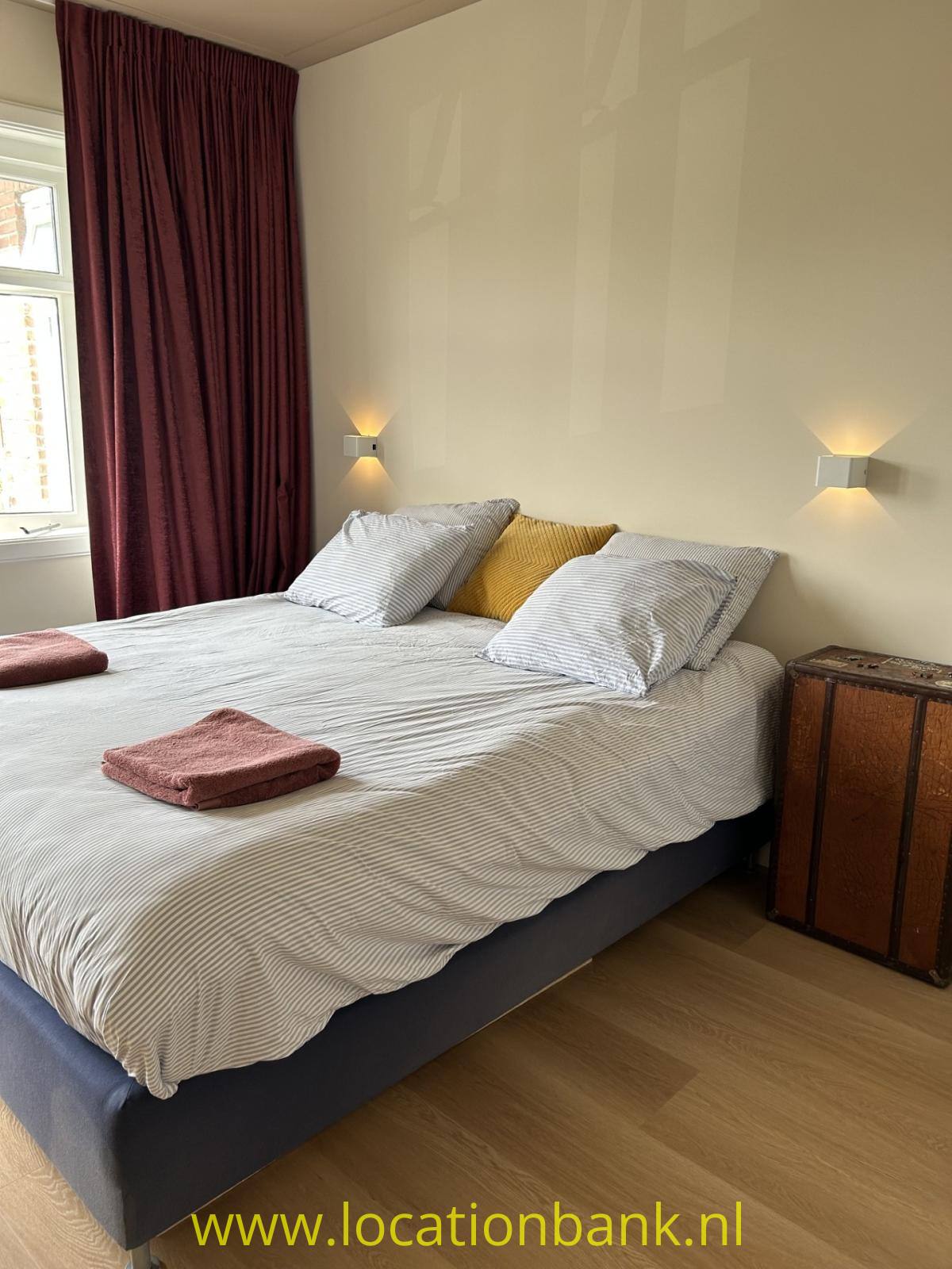 Ein Doppelbett mit gestreifter Bettwäsche, mehreren Kissen und gefalteten Handtüchern steht neben einem Holzschrank.