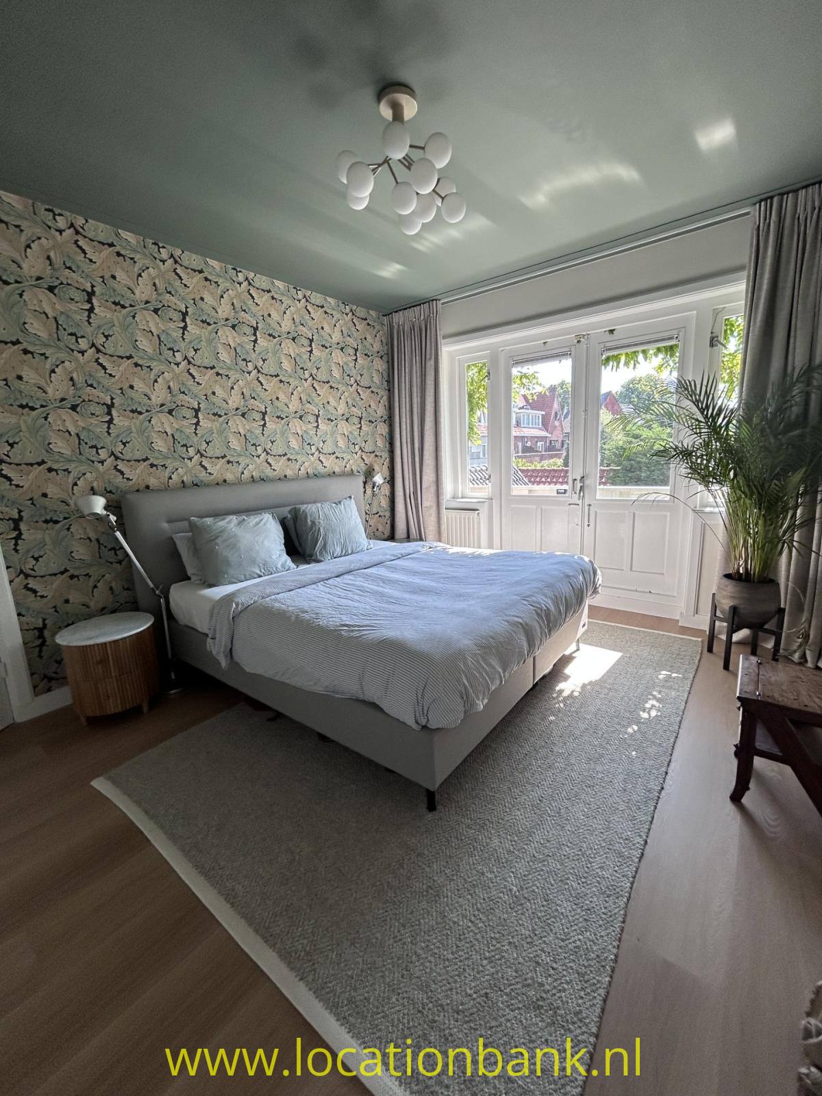 Schlafzimmer mit einem bezogenen Bett, botanischer Tapete, hohen Fenstern, Vorhängen und einer großen Zimmerpflanze.