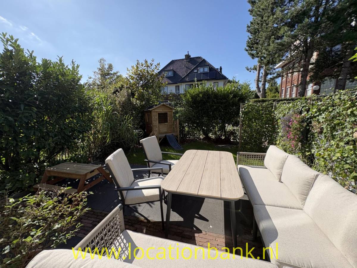 Terrasse mit einem langen Gartentisch, Stühlen und einem Ecksofa, umgeben von grünen Hecken und Bäumen.