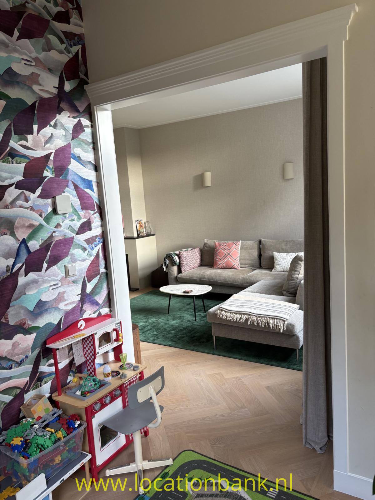 Durch eine Türöffnung ist ein Wohnzimmer mit grauem Ecksofa und grünem Teppich sichtbar.