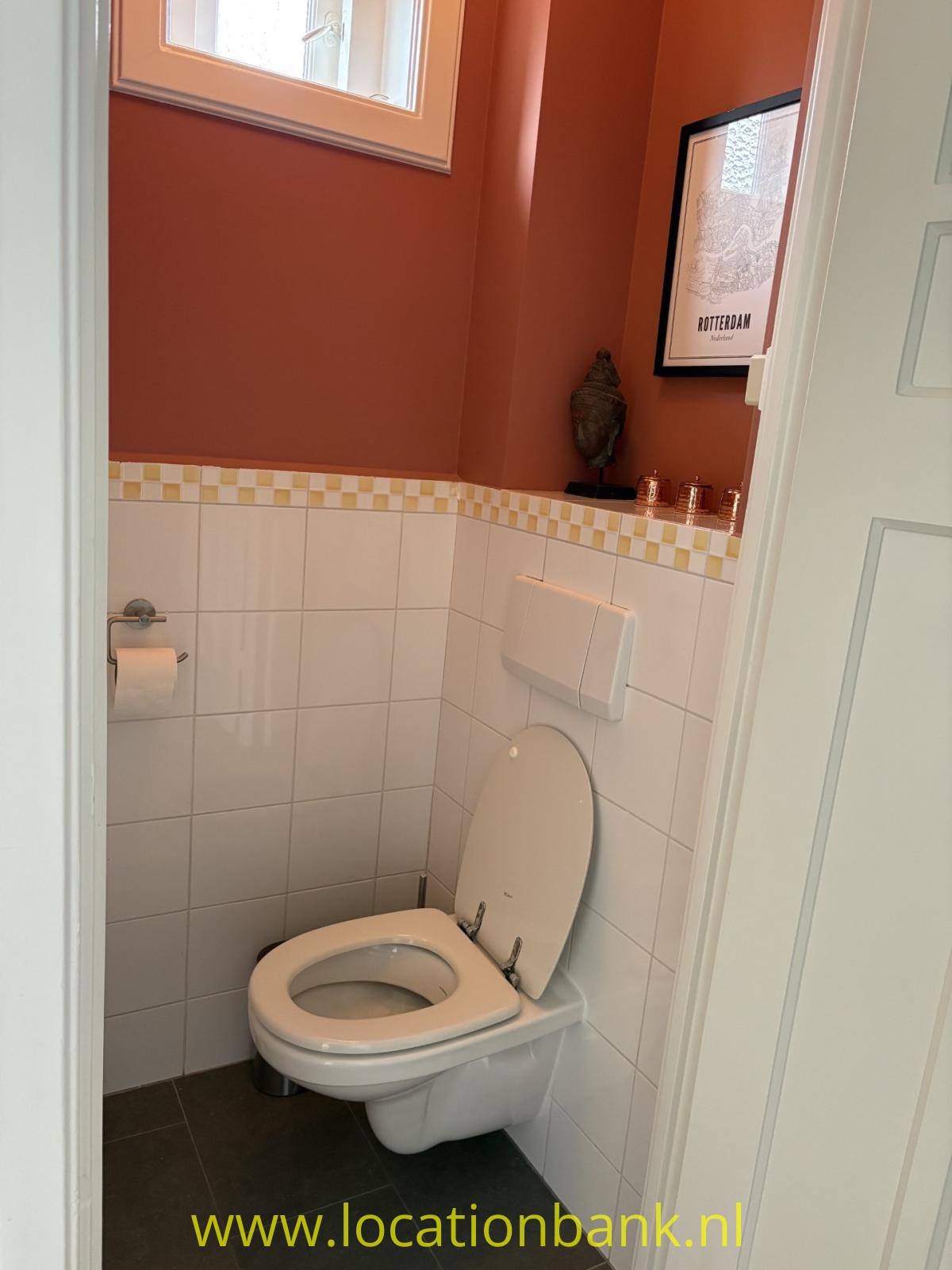 WC mit weiß gefliesten Wänden, einer dekorativen Fliesenbordüre, dunklen Bodenfliesen und terrakottafarbenen Wänden.