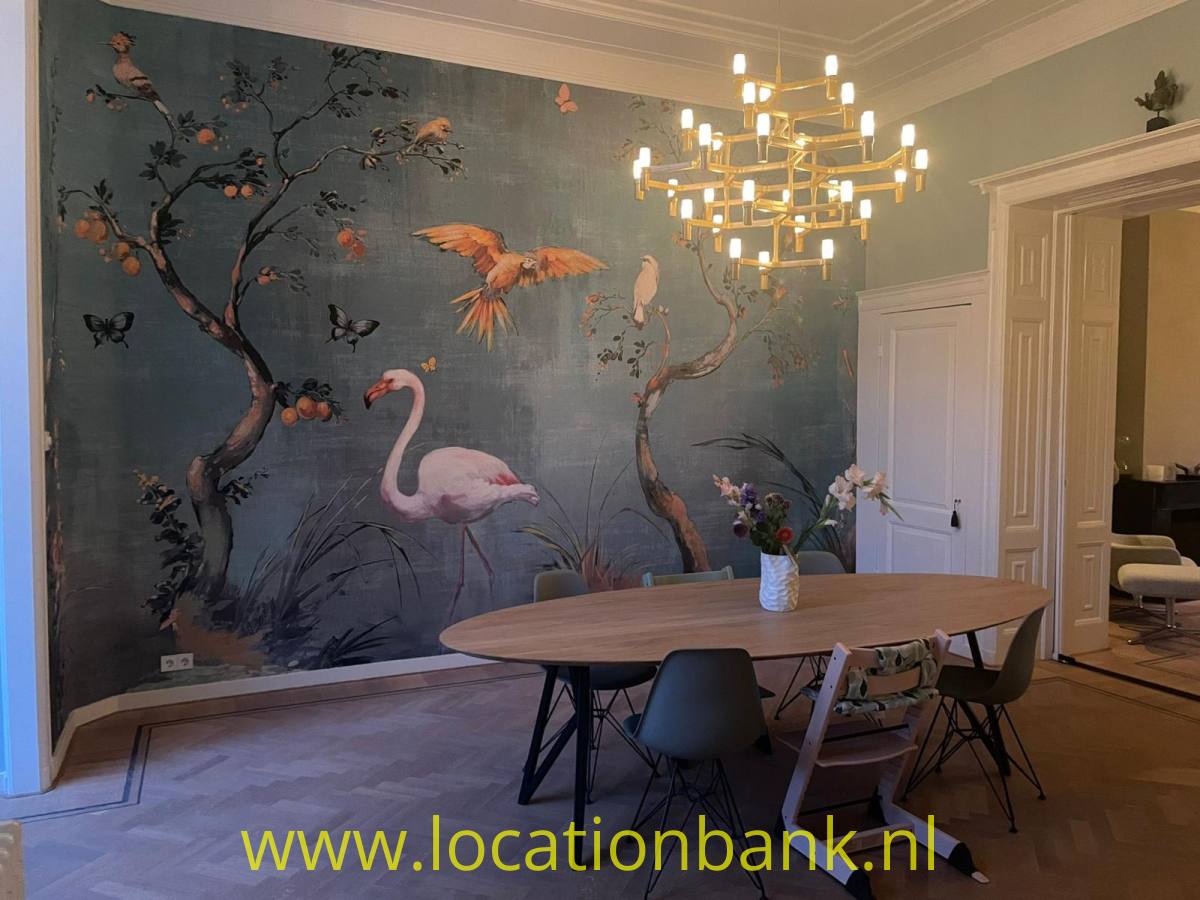 Raum mit einem Wandgemälde von Flamingos, Vögeln und blühenden Zweigen, einem ovalen Tisch und einem Kronleuchter.