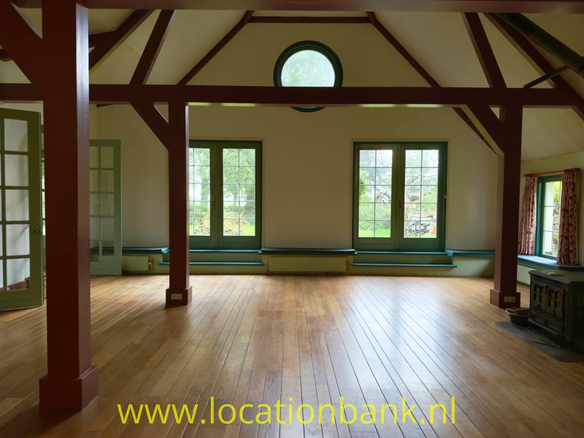 Geräumiger Raum mit sichtbaren dunklen Holzbalken, einem Holzboden, großen Fenstern und einem runden Fenster im oberen Wandbereich.