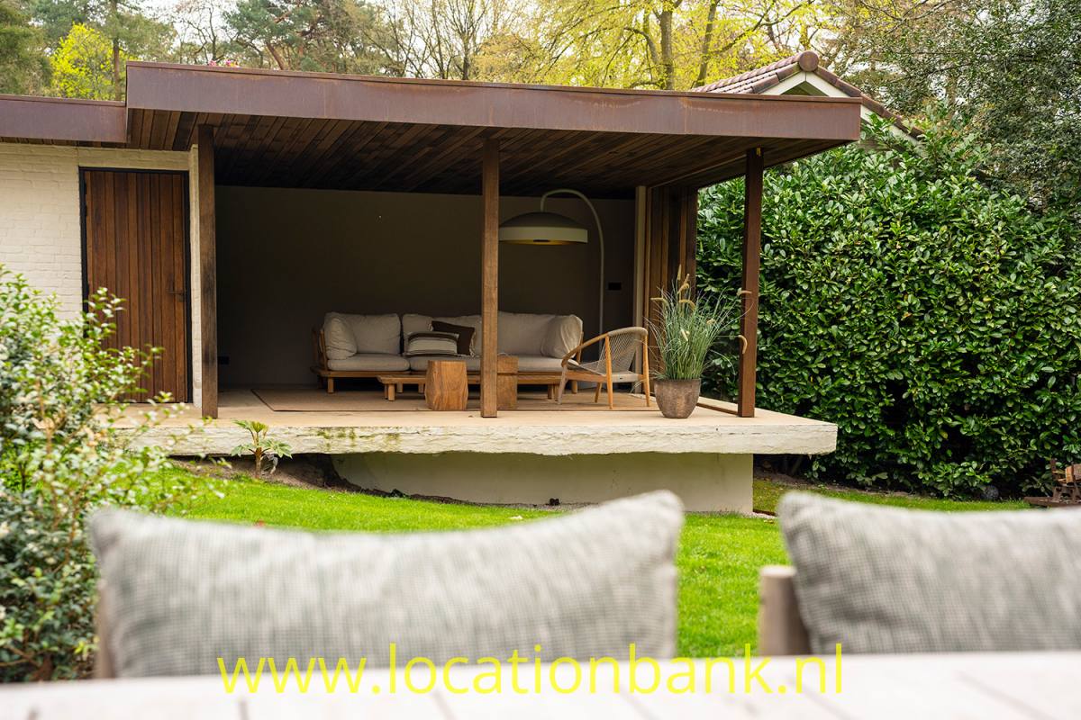 Eine überdachte Holzterrasse mit Sitzmöbeln befindet sich erhöht zwischen Pflanzen; im Vordergrund liegen Kissen.