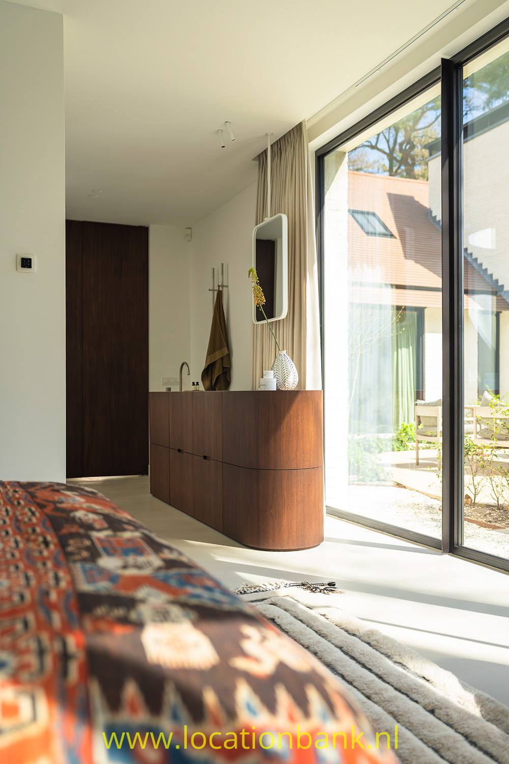 Abgerundetes Sideboard aus dunklem Holz neben hohen gläsernen Schiebetüren, mit farbenfrohem gemustertem Stoff im Vordergrund.