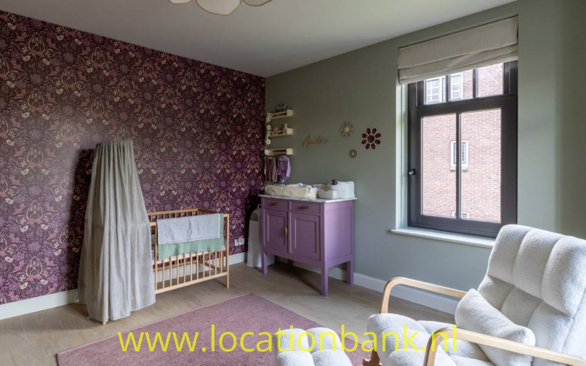 Zimmer mit dunkelvioletter Blumenmustertapete, einem Holz-Kinderbett, einer Wickelkommode, einem Sessel und einem großen Fenster.