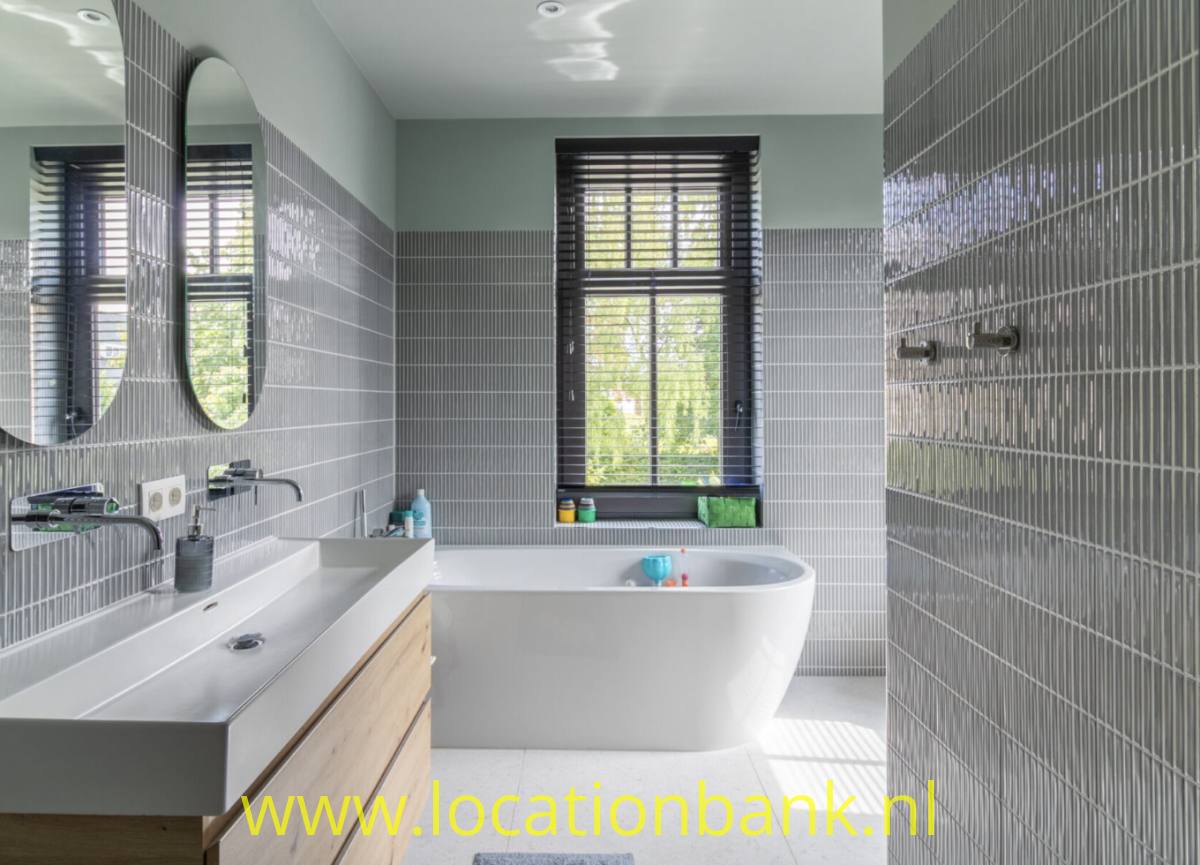 Freistehende weiße Badewanne in einem Badezimmer mit grauen Wandfliesen, zwei ovalen Spiegeln und Fenstern mit Jalousien.