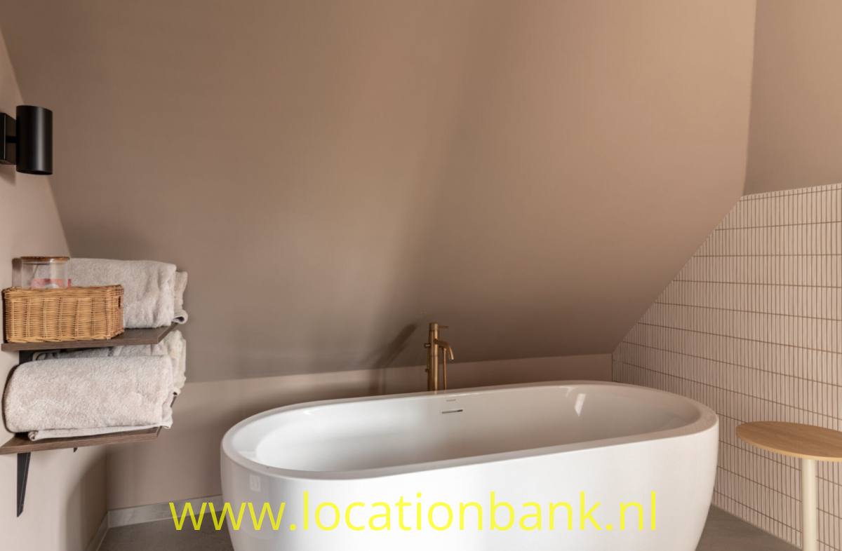 Freistehende weiße Badewanne unter einer Dachschräge, daneben gerollte Handtücher und ein Holzhocker.