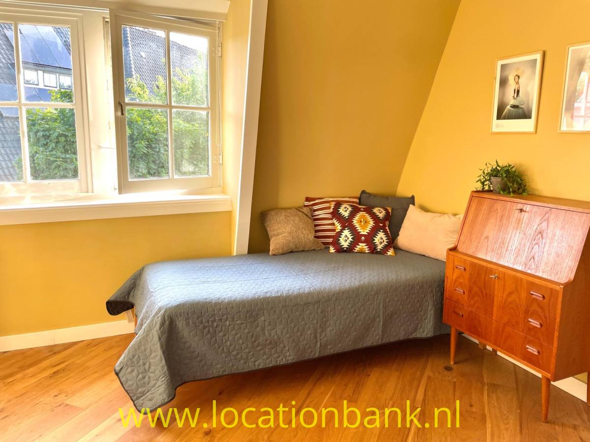 Ein Bett mit einer grauen Tagesdecke und gemusterten Kissen steht neben einer Holzkommode in einem gelben Raum.