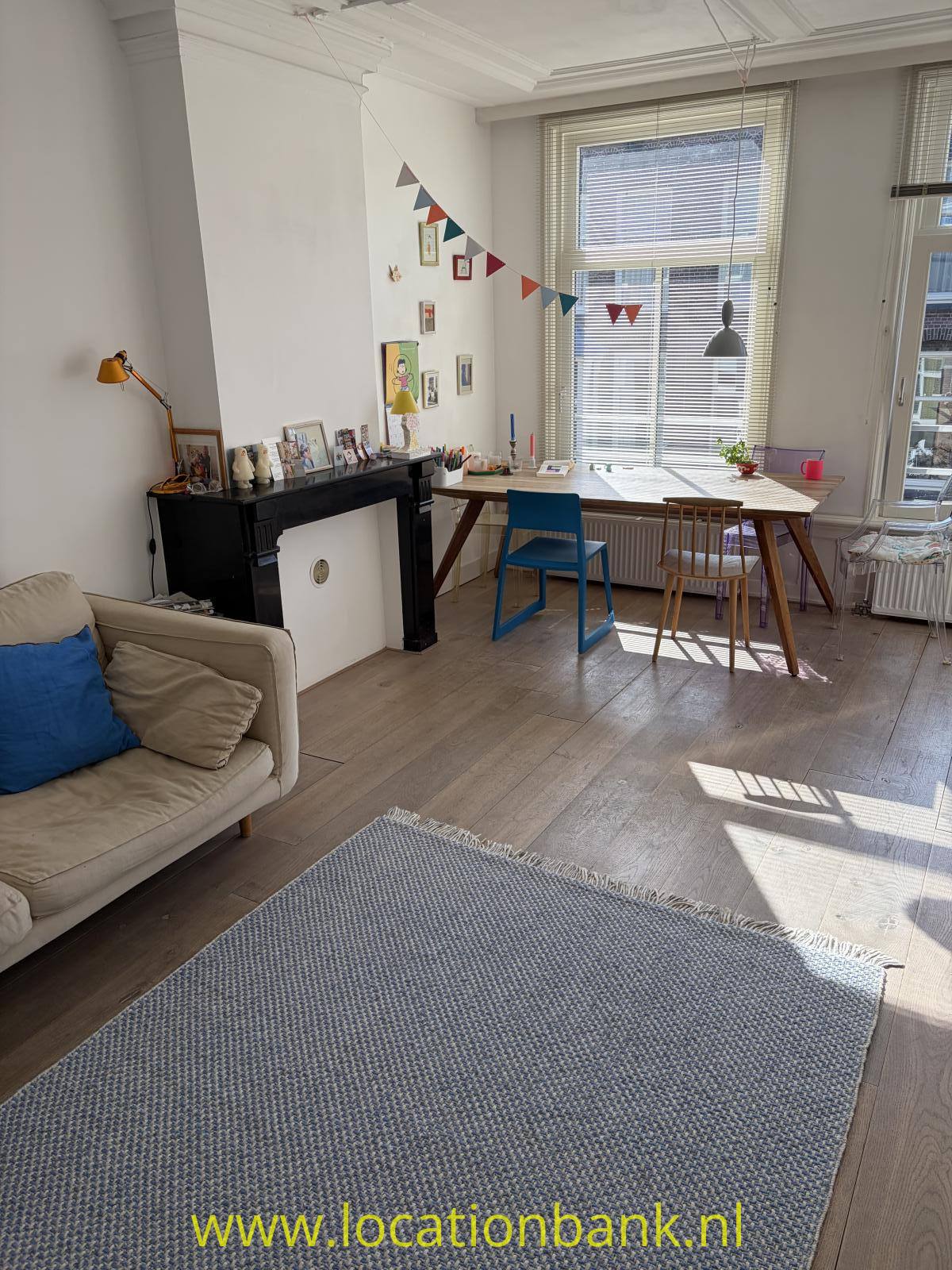 Wohnzimmer mit einem großen Fenster mit Jalousien, einem Schreibtisch, zwei Stühlen, einem Sofa und einer schwarzen Kaminverkleidung.