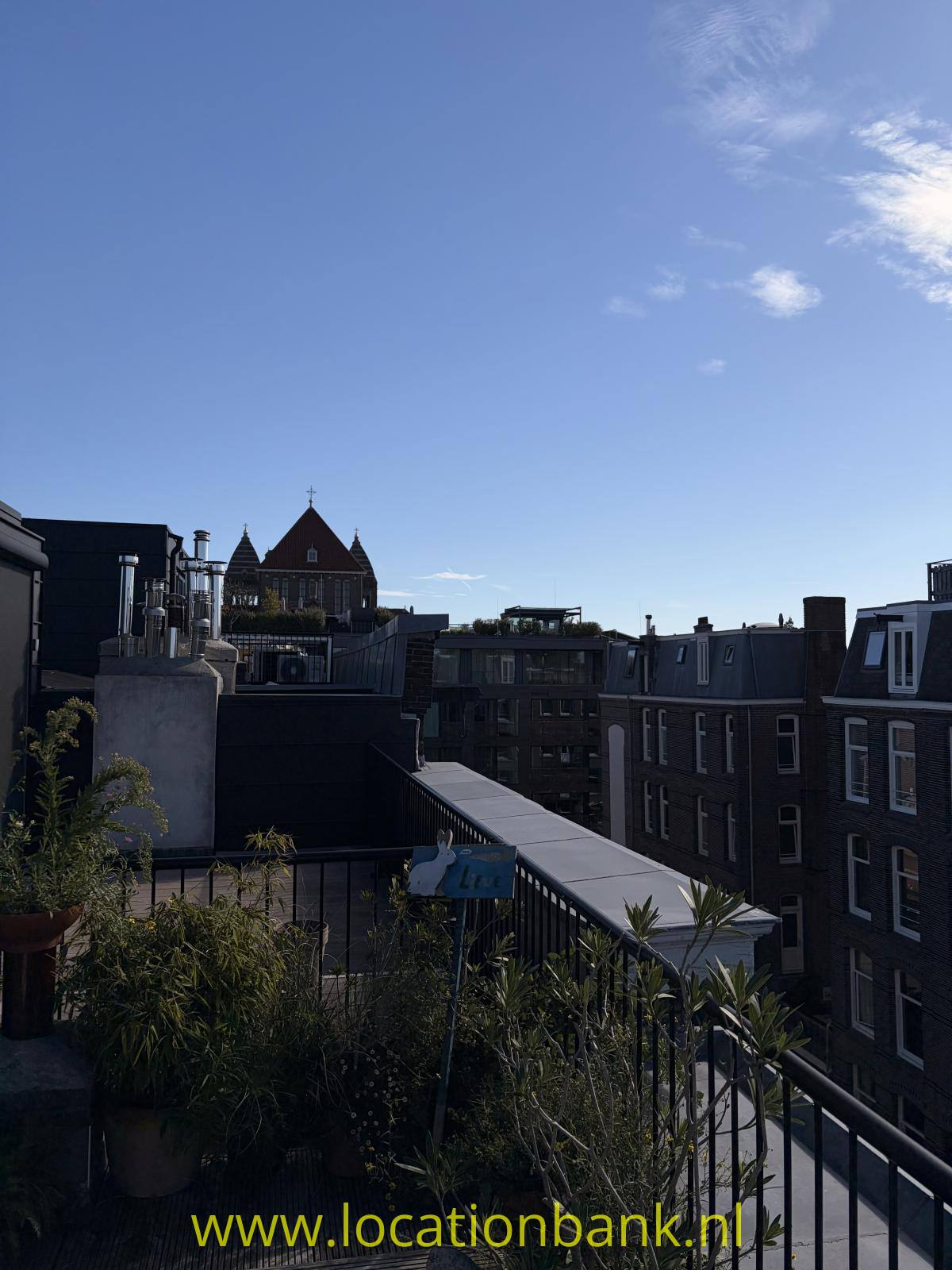 Dachterrasse mit Pflanzkübeln und Metallgeländer, umgeben von Backsteingebäuden und einem spitzen Turm im Hintergrund.