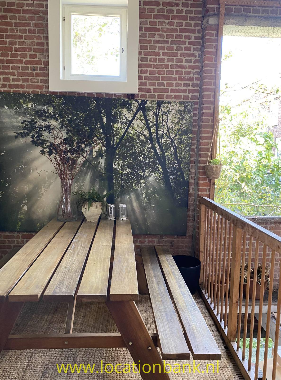 Ein hölzerner Picknicktisch mit Bänken steht vor einer Backsteinwand neben einem Holzgeländer.