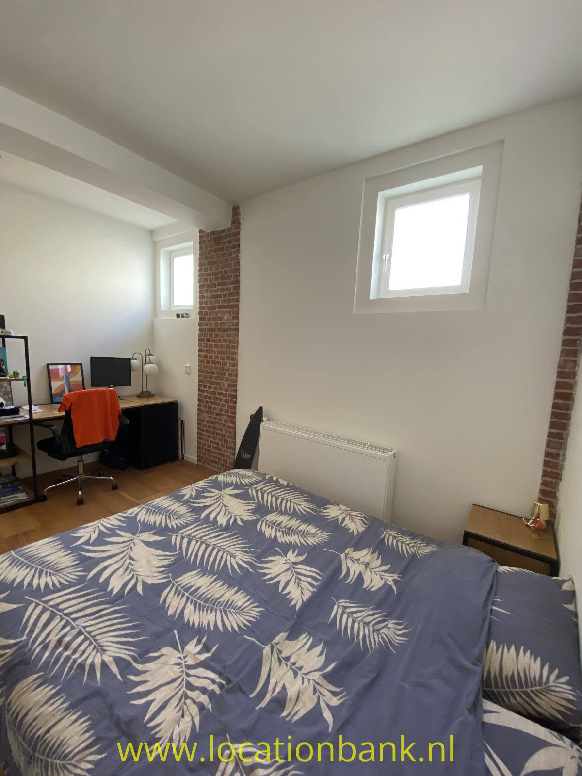 Schlafzimmer mit einem Bett mit blauer Bettwäsche mit Blättermuster, einem Schreibtisch und freiliegendem Ziegelmauerwerk.