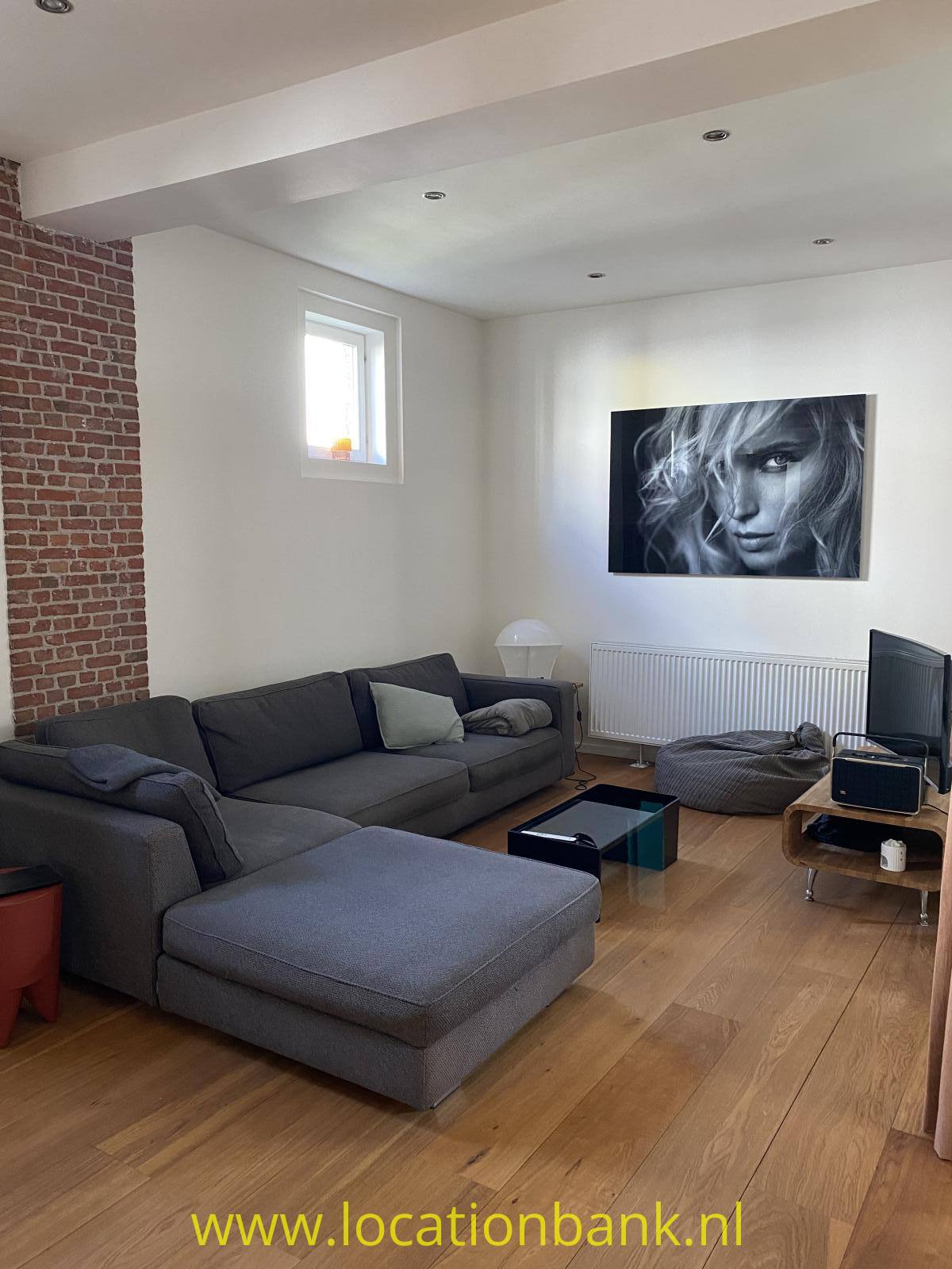 Graues Ecksofa mit Chaiselongue in einem Wohnzimmer mit Holzboden, Backsteinwand und Schwarz-Weiß-Porträt.